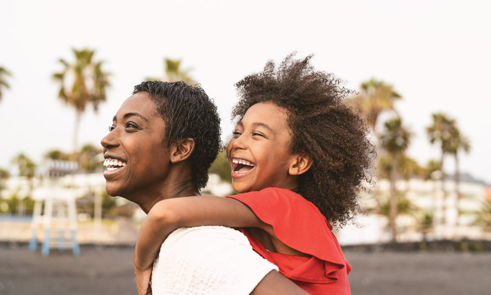 Mom giving her daughter a piggyback ride on the beach while laughing