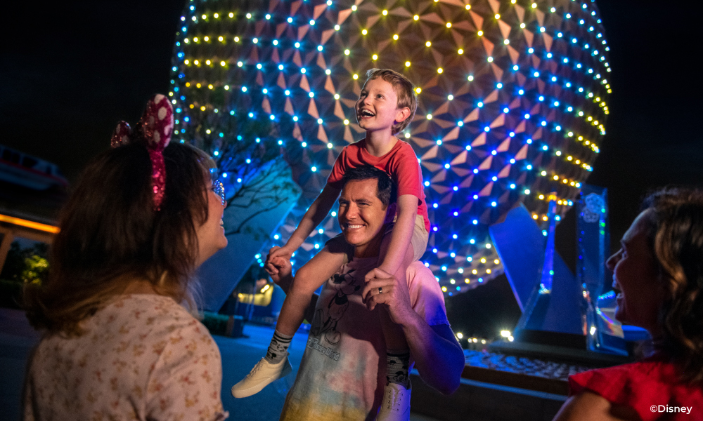 Son riding dad's shoulders at night in front of the illuminated EPCOT ball