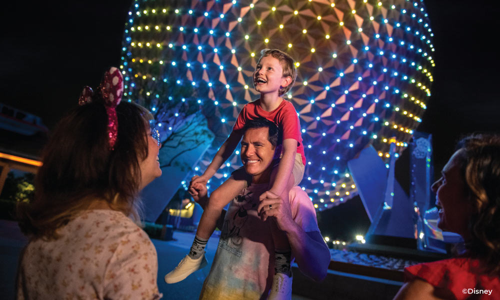 Young boy on his dad's shoulders at night with the illumicnated EPCOT Ball behind them