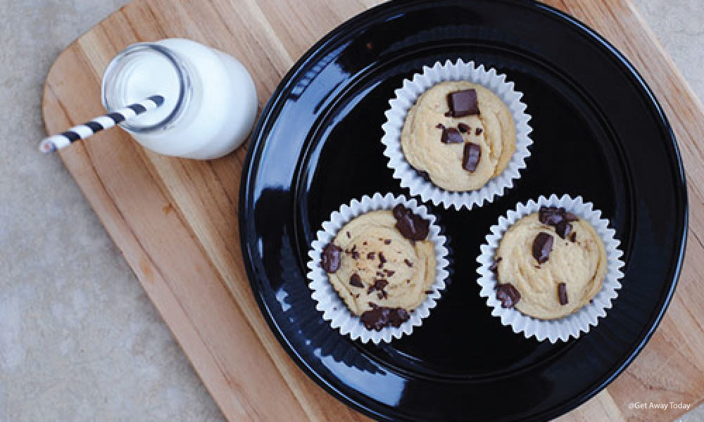 Three chocolate chip cookies baked in white baking cups on a black plate with a cup of milk